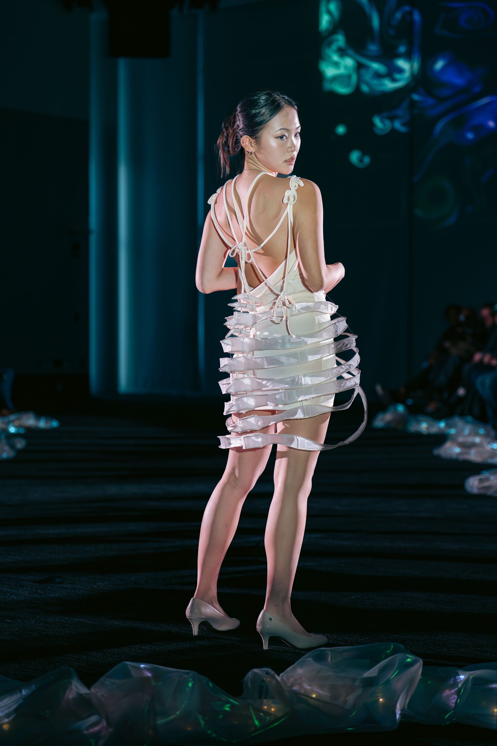 Model walks down the runway in a sculptural white dress with an open back, tied shoulder straps, and a ribbed cage-like skirt made of curved translucent strips. The audience is dimly visible in the background.